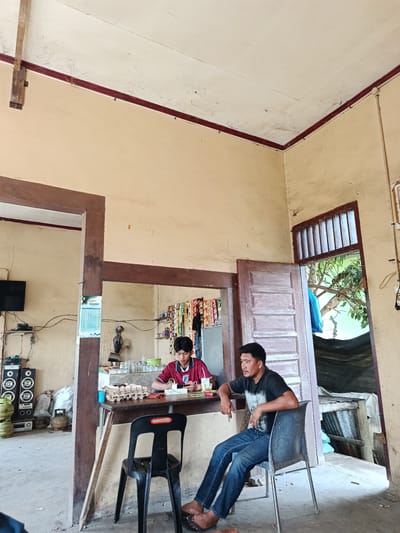 This image depicts an indoor scene at a small establishment, possibly a shop or eatery, located in Lancok, Indonesia. In the foreground, a man with dark hair and a mustache, wearing a black t-shirt and blue jeans, is seated in a dark plastic chair. He is looking to his left. Behind him, a young man with dark hair, wearing a red and white striped polo shirt, is seated at a counter. He appears to be using a mobile phone and has a drink in front of him. The counter displays a carton of eggs and various items behind it, including packaged goods. The environment appears somewhat rustic, with textured yellow walls and wooden architectural elements. To the left of the counter, there are speakers and gas cylinders. A doorway to the right reveals an outdoor area with trees and corrugated metal structures. The overall atmosphere suggests a casual, everyday setting.
