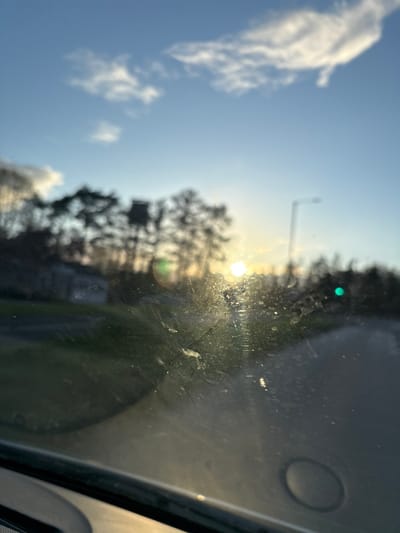 The image shows a view through a car windshield, oriented sideways. The sun is visible through the trees and possibly buildings, creating a bright glare. To the left of the sun, a few clouds are visible in a blue sky. A street light post can be seen in the mid-ground. The windshield is dirty and has smudges, affecting visibility. A green light is also visible above the trees. The dashboard of the car is visible in the bottom right of the frame. The location cannot be determined based on the image alone.