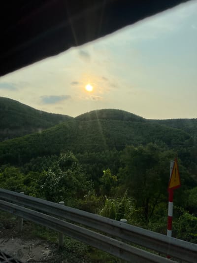 The image, taken from a moving vehicle, depicts a mountainous, forested landscape in Aê Thi, Vietnam. The sun is visible in the sky, partially obscured by clouds. A yellow and red road sign is positioned on the right side of the frame, near a metal guardrail. The guardrail is in the foreground, suggesting the image was taken from a road or highway. The mountain range extends into the distance, covered in dense vegetation. A portion of the vehicle's interior is visible on the left side of the frame.
