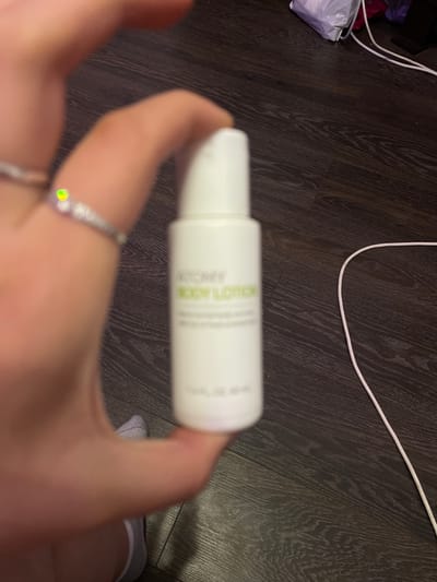 A close-up shot shows a hand holding a small white bottle of body lotion. The bottle has green text that is partially legible, but appears to say "BODY LOTION". A portion of a ring with a small, iridescent green and yellow stone is visible on one of the fingers. The background is a dark wooden floor. A white cable snakes across the floor in the upper right corner of the image, and some white and purple packaging is visible further in the background. The scene is not identifiable as being in Shcheglovo, Russia.