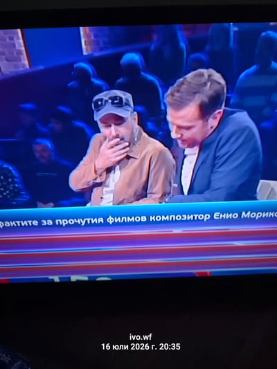 The image shows a close-up, slightly blurry, full-frame shot of a television screen. The screen displays two men seated at a table, with other people blurred in the background. The man on the left is wearing a light brown jacket over a white shirt and a white baseball cap with dark sunglasses. He is covering his mouth with his hand, appearing to be in distress or surprise. The man on the right is wearing a dark blue suit with a white shirt and a light-colored tie. He is looking down at his hands, which are resting on the table in front of him. A blue banner with white text is visible across the bottom of the screen, partially obscuring the lower bodies of the men. The text on the banner appears to be in Cyrillic script. Below the television screen, in the lower portion of the image, there is blackness and some indistinct text, including "ivo.wf" and "16 юли 2026 г. 20:35". The overall scene suggests a televised program or broadcast. No specific location in Chal, Bulgaria is discernible from the image content.