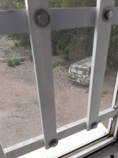 An outdoor, ground-level view looks through a white barred window and shows a camouflaged van parked on a gravel and dirt lot. The van is facing away from the viewer and to the right. The camouflaged pattern on the van consists of irregular shapes in shades of green and grey. The van has two windows on its rear door, both appearing to be covered or tinted. Red taillights are visible on either side of the rear of the van. The ground is composed of light brown dirt and small grey gravel, with sparse patches of green vegetation. In the background, several trees with green foliage are visible. The overall scene suggests a somewhat unkempt or rural environment. This image was taken in the city of Satbayev, Kazakhstan.