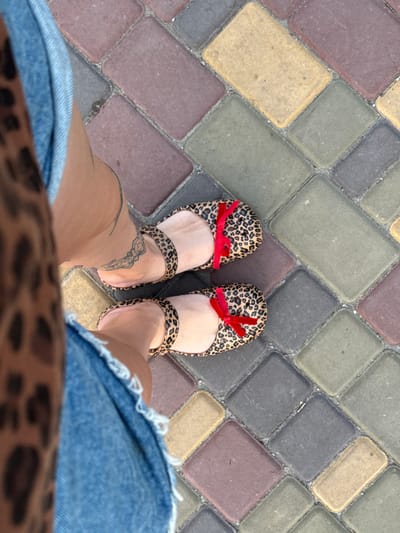The image shows a person's feet wearing leopard print ballet flats with red bows, standing on a cobblestone path. The cobblestones are of various colors including red, yellow, green, and gray. The person is wearing denim shorts and a leopard print skirt. The scene appears to be outdoors, possibly in a city like Piatykhatky, Ukraine, given the context of the prompt.