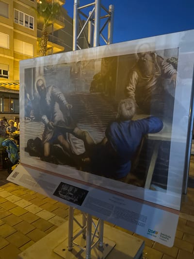 The image shows a large framed artwork displayed outdoors in Mazarrón, Spain, under a twilight sky. The artwork is a reproduction of a painting featuring several figures. In the foreground of the painting, a man with a beard, wearing a white robe, is pulling a younger man, who is lying down and wearing a blue tunic, by the legs. Another figure, with a beard and wearing a brown robe, is visible in the background. The painting depicts a scene that appears to be biblical or historical, with dramatic lighting and a subdued color palette.
The artwork is mounted on a metal stand and is positioned on a tiled walkway. To the left of the artwork, there are tables and chairs set up, suggesting an outdoor seating area for a cafe or restaurant. Some people are seated at the tables, and a palm tree is visible behind them, adjacent to a building with multiple balconies and windows. The building appears to be residential or commercial, with warm lighting emanating from some of the windows.
At the bottom of the artwork, a descriptive panel provides information about the painting and its artist. Text visible on this panel includes "TINTORETTO" and "125 años luz Iberdrola España Fundación". A QR code is also present on the panel. The overall scene captures an art exhibition or display integrated into the public space of the city.