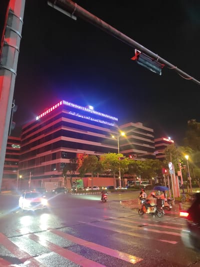 The image depicts a nighttime street scene in Nanhai District, China, likely at an intersection. A multi-story building with illuminated signage reading "Global Hardware & Building Materials Manufacturing Base" and in Chinese and Arabic characters dominates the background. Red and blue neon lights highlight the building's facade. Several vehicles, including cars and motorbikes, are present on the wet street, suggesting recent rain. Three individuals are riding on motorbikes, one of whom is holding a blue umbrella. Traffic lights are visible and illuminated. The street is marked with crosswalks, and streetlights cast yellow illumination. The overall atmosphere is urban and active, despite the late hour and wet conditions.