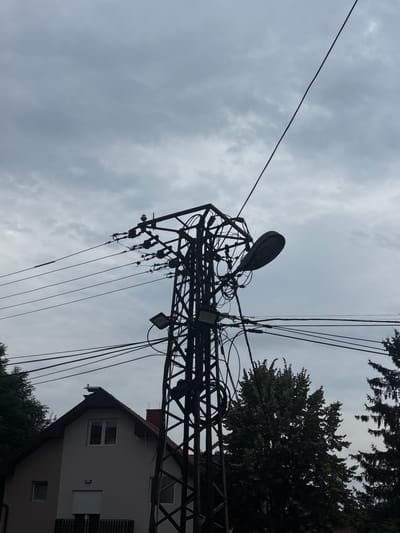 The image depicts an overcast sky over a residential area in Loznica, Serbia. A tall, lattice-style electrical pylon dominates the center of the frame, heavily laden with numerous wires and cables. A street lamp is attached to the pylon. To the left of the pylon, a portion of a light-colored house with a dark-tiled roof is visible. The house has at least two windows and a retractable shutter on one of them. Trees, some appearing to be evergreens, are scattered around the scene, with one dense cluster of foliage on the right side of the image. The overall impression is one of a typical, somewhat cluttered, utility infrastructure setup in a suburban or semi-urban environment.