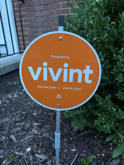 A round, orange Vivint security system sign is planted in the ground in front of a brick building with a black railing and a window. A green bush is to the right of the sign. The sign has "Protected by vivint" written at the top in white text, followed by the Vivint logo and the text "855.484.8468 | vivint.com". The sign is wet, indicating recent rain. The ground in front of the sign is covered in mulch. The image is a medium shot, taken from a slightly elevated angle.