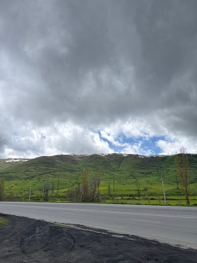 A landscape image depicts a winding road adjacent to a verdant hillside under a cloudy sky, possibly located near Tsovagyugh, Armenia. The hillside exhibits a steep gradient, marked by terraced fields and scattered trees. A roadway separates the hillside from the asphalt road and has power poles that run in a linear fashion. The road itself is gray and stretches away and shows road markings. The sky is filled with dark gray clouds, interspersed with patches of bright blue, indicating varying weather conditions. The overall composition is one of natural beauty juxtaposed with infrastructure, suggesting a rural or semi-rural setting.