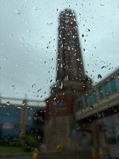 The image is a close-up, blurred view of raindrops on a window, looking out onto a city street scene. The background is out of focus, showing a tall, brick structure that appears to be a chimney or tower, possibly part of an industrial building, situated next to a lighter-colored building with teal-colored windows. The sky is overcast and gray. The foreground is dominated by numerous water droplets of varying sizes, clinging to the glass surface, distorting the view of the outside world. The scene suggests a rainy day in an urban environment, possibly in the Trafford area of the United Kingdom, though specific landmarks are not identifiable due to the blur and rain. No people or specific actions are discernible.