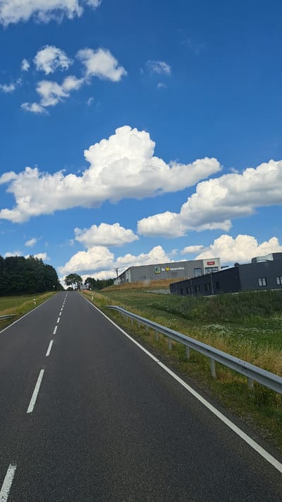 The image depicts a view from a moving vehicle on a two-lane asphalt road in Höhn, Germany. The road is marked with white dashed lines on the left lane and a solid white line on the right edge, indicating a clear path forward. A metal guardrail runs along the right side of the road, separating it from a grassy verge dotted with wildflowers.

In the background, to the right of the road, are several modern industrial buildings. One building prominently displays the yellow "E" logo of an Edeka supermarket chain, with the text "Wir leben Lebensmittel" (We live groceries) written beside it. Another building to its right is dark grey and appears to be a warehouse or commercial property.

The landscape to the left of the road is characterized by a dense line of green trees, forming a forest edge. Beyond the trees, the sky is a vibrant blue, filled with large, fluffy white cumulus clouds. The overall scene suggests a sunny day in a rural or semi-industrial area outside of the main town center of Höhn. There are no people or animals visible in the frame.