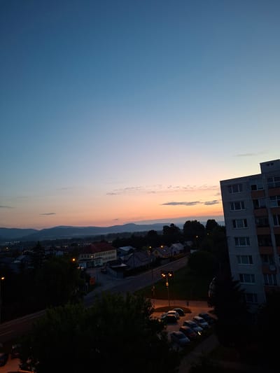 This is a high-angle, long shot photograph of a city scene at dusk, likely in Ilava, Slovakia. The sky displays a gradient of colors, transitioning from a deep blue at the top to lighter blues and oranges near the horizon, with some scattered clouds. In the foreground, on the right side, is the side of a multi-story apartment building with numerous windows. Below and to the left of the building, a street is visible, lined with houses and trees. Several parked cars are on the street and in a parking lot. In the distance, there are rolling hills or mountains. The lighting suggests it is early morning or late evening.