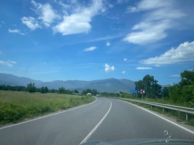 The image captures a view from inside a vehicle traveling on a paved road that curves to the left, heading towards rolling, mist-covered mountains under a bright blue sky dotted with white clouds. The road has a white dashed line down the center and a white solid line on the right edge, bordered by a metal guardrail. To the left of the road, there is a grassy field with some scattered trees. To the right, dense green foliage and trees line the road. A blue road sign with white lettering indicating "Berkovitsa 2" and "Berkovitsa" in Bulgarian and English, respectively, is visible on the right side of the road, accompanied by a directional arrow. Further down the road, two vehicles are visible in the distance, and the hood of the car from which the photo is taken, featuring a Mercedes-Benz emblem, is in the foreground. The scene depicts a rural, mountainous landscape in the vicinity of Mezdreya, Bulgaria, on a clear day.