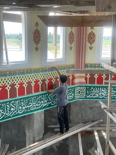 An interior shot depicts a male individual meticulously applying paint or decorative elements to a wall within a building under construction or renovation, identified as being in Mns Asan, Indonesia. The man stands barefoot on a makeshift wooden scaffolding plank. He wears a paint-splattered grey t-shirt and dark trousers, with his right hand extended upwards, holding a small tool (likely a brush) against a red section of the wall. His left hand rests flat against the wall below.
The walls are elaborately decorated with Islamic motifs. The upper wall features a light yellow/cream background with symmetrical pink and green floral/geometric patterns, flanked by tall, arched windows with white frames. Below this, a multi-colored geometric frieze sits atop a red band adorned with a repeating stylized arch pattern. Further down, a prominent dark green band displays intricate white Arabic calligraphy. A square geometric lattice pattern is visible within this green band to the right. A pinkish-orange fluted pillar or corner detail is present on the right side of the frame. Rough wooden beams of an unfinished ceiling or roof structure are visible at the top. The area below the scaffolding consists of unfinished grey concrete.
Through the windows, a rural Indonesian landscape is visible, featuring expansive, water-filled rice paddies reflecting the sky, interspersed with palm trees, and a distant utility pole.