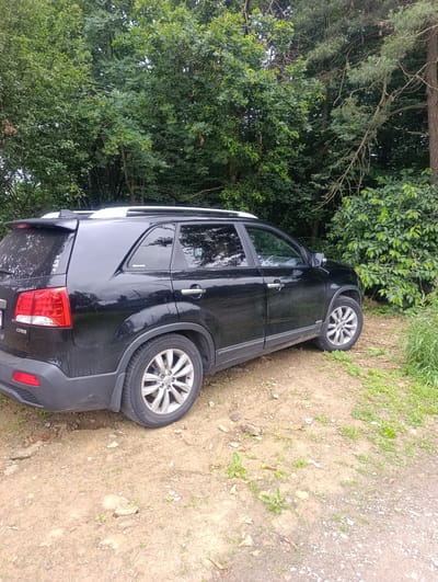 The image shows a black Kia Sorento parked on a dirt path surrounded by lush green trees. The car is positioned at an angle, with the rear and side visible. The path appears to be unpaved and somewhat uneven, with small stones and sparse vegetation. The trees form a dense backdrop, suggesting a natural or wooded environment. There are no people visible in the image. The scene is likely a rural or semi-rural location, potentially on the outskirts of Myślenice, Poland, given the vegetation and unpaved road conditions often found in such areas.