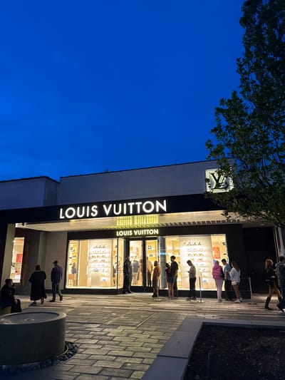 The image depicts a Louis Vuitton store at night in Oak Brook, Illinois. The illuminated facade of the store features the "LOUIS VUITTON" name vertically in white, prominent above the entrance. People are present near the storefront, some looking into the display windows. The sky is a deep blue, with the outlines of tree branches visible above the building. The sidewalk in front of the store is paved with rectangular stones. There is a large, cylindrical concrete object along the edge of the sidewalk near the bottom of the image.