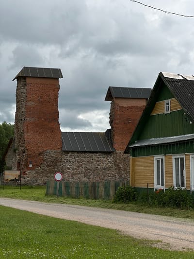 The image shows the remains of a brick tower and a partially intact stone wall, possibly part of a historical ruin or fortification. Above the ruins, a section of a building with a green wooden roof and yellow siding is visible, featuring windows and a small attic window. The building appears to be modern and contrasts with the weathered look of the ruins.

A gravel path runs diagonally across the right side of the image, bordered by a well-maintained green lawn with scattered small white flowers. A dark green wooden fence runs along the stone wall, separating it from the path and lawn.

The sky is overcast with heavy, dark clouds, suggesting a potentially stormy or dull weather condition. A single black wire is visible in the upper left corner of the frame, extending out of view. A circular sign with a red border and a white interior, likely a traffic or informational sign, is mounted on the stone wall.

Below the stone wall, to the left, a wooden information board is partially visible, with what appears to be a map or historical depiction. The overall scene suggests an outdoor location, possibly a park or historical site, under a cloudy sky. There is no clear indication of people or specific activities taking place, other than the presence of a path suggesting pedestrian access. The combination of historical ruins and a more modern building creates a layered historical and visual context.