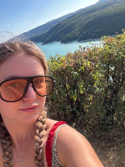 A young woman with blonde hair in two braids and wearing large, dark-rimmed sunglasses with orange lenses takes a selfie. She is wearing a red top with a patterned strap. A gold necklace with a cross pendant is visible. Behind her, a bright turquoise lake is nestled between tree-covered mountains under a clear blue sky. A dense hedge of green and brown foliage is in the foreground, obscuring some of the view. The image appears to have been taken on a sunny day. The location is Moirans-en-Montagne, France.