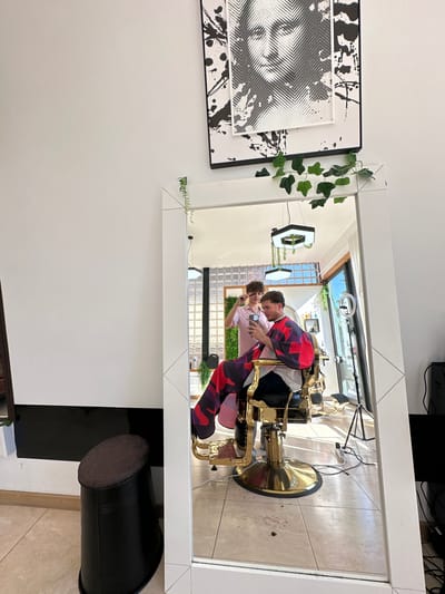 A reflected scene in a mirror shows two individuals in a barber shop in Celorico de Basto, Portugal. One person, wearing a red and purple patterned jacket, is taking a picture with a phone. The other person wears a light pink shirt. The reflection also shows a gold-plated barber chair, decorative plants, a black-and-white abstract wall art, a light-colored tiled floor, and lighting equipment.