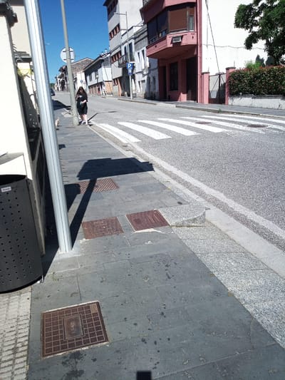 The image depicts a street scene in Castellfollit de la Roca, Spain. A young woman with dark hair, wearing a black t-shirt and shorts, stands on the sidewalk next to a lamppost, looking at her phone. She is positioned near a pedestrian crossing. To her left, a gray trash receptacle with visible holes is situated on the sidewalk. The sidewalk is paved with dark gray tiles, and several square metal manhole covers are embedded in it. A shadow from the lamppost falls across the sidewalk. Across the street, there are buildings with white and pink facades. The street is asphalted, and the pedestrian crossing is marked with white stripes. The sky is clear and blue.