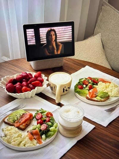 This media file captures an inviting breakfast or brunch scene on a polished wooden table, set indoors, possibly in Ros Na Rí, Ireland. Two white, oval plates are laid out on linen placemats, each featuring a balanced meal. Both plates contain fluffy scrambled eggs, a fresh green salad with lettuce, diced cucumber, and halved cherry tomatoes, and a slice of toast topped with mashed avocado, smoked salmon, and a sprinkle of sesame seeds, possibly accompanied by other seeds.

Between the main plates, a white, leaf-shaped bowl is filled with vibrant red strawberries. Two clear glasses hold a frothy, light-colored beverage, likely lattes or cappuccinos, indicating a leisurely morning. A distinctive small, light-colored wooden cup or mug, featuring a stylized letter "R" or "Я" on its side, is also present.

On the left side of the table, a tablet is propped up, displaying a video of a woman with dark hair, looking off-screen. She appears to be wearing a dark top and a ring on her right hand, with vertical window blinds visible in the background of her video. In the soft background of the main image, light-colored upholstered furniture and cushions are visible, suggesting a comfortable and relaxed home environment. The natural lighting suggests it is daytime. The overall ambiance is calm and pleasant, set for a shared meal.
