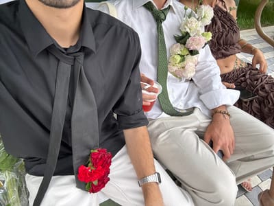 An eye-level shot captures three individuals seated outdoors in Nor Hachin, Armenia. The person on the left, a male, wears a black collared shirt with rolled sleeves, an untied black tie, and white pants. A cluster of red flowers is tucked into his shirt fabric near his left hip. He wears a silver watch on his left wrist. The central figure, a male, wears a white collared shirt with rolled sleeves, a green patterned tie, and light-colored pants. A floral arrangement of white and light pink flowers is pinned to the right side of his shirt. He holds a clear plastic cup containing a reddish beverage in his left hand and has a brown beaded bracelet with a silver cross on his right wrist. The person on the right, a female, wears a brown ruffled crop top and a matching long skirt, with green beaded body jewelry visible on her torso. A smartphone rests on her lap. The background shows blurred greenery, and paved ground is visible in the lower right foreground.