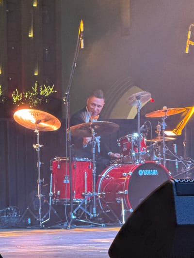 The image, rotated 90 degrees clockwise from its correct orientation, captures a male drummer performing on a stage. The central focus is a vibrant red drum kit and the drummer behind it.
The drum kit features several components: a prominent large red bass drum with the "YAMAHA" logo clearly visible in white lettering on its black head, several smaller red drums (likely snare and tom drums), and multiple cymbals. One of the cymbals, positioned in the lower left foreground (after rotation), reflects a bright light source and has the letters "HH" visible, indicating a Zildjian Hand Hammered cymbal. Another cymbal in the upper center also reflects light strongly. Microphones on stands are strategically placed around the drums to capture sound. In the upper left corner, part of a brass instrument, possibly a trumpet or trombone, can be seen. A large black speaker is visible in the upper right corner of the frame, partially obscuring the view of the stage.
The drummer is a man with short, dark hair, looking towards the viewer with a slight smile, suggesting enjoyment or acknowledgement of the audience. He appears to be wearing a dark suit jacket over a lighter-colored shirt. His hands are near the drums and cymbals, indicating he is actively engaged in playing or has just finished a segment.
The setting appears to be a live performance venue. The background is dark and out of focus, typical of stage lighting that highlights the performers. Bright stage lights illuminate the drum kit, creating reflections on the shiny surfaces of the drums and cymbals. In the lower left background, some blurry, warm lights can be seen, possibly part of the stage decor or audience area lighting. The photo seems to be taken from an elevated position, looking down slightly onto the stage.