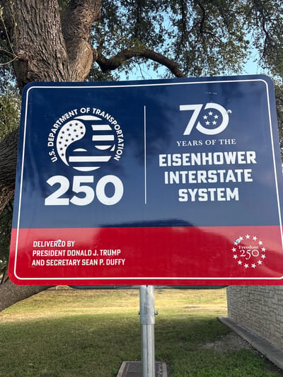 A blue and red sign commemorating "75 Years of the Eisenhower Interstate System" is displayed on a metal pole in Sonora, United States. The sign features the U.S. Department of Transportation logo, the number "250", and text indicating it was "Delivered by President Donald J. Trump and Secretary Sean P. Duffy." A small circular emblem with stars and the text "Freedom 250" is also visible on the red section of the sign. The sign is positioned outdoors with a large tree and green grass in the background.