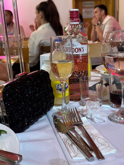 The scene is set indoors at a social gathering or restaurant in Debrashtitsa, Bulgaria, under ambient pink and white artificial lighting. The foreground features a white tablecloth with various dining and drinking items. A black, heavily beaded or sequined clutch purse with a silver clasp rests on the left. To its right, a tall wine glass contains a clear, yellowish liquid, possibly white wine. A yellow Fanta bottle is partially visible behind this glass. Centrally located is a bottle of Gordon's Pink Gin with a red cap and label depicting strawberries. Further right, an empty clear glass tumbler and a larger wine glass containing a dark liquid (possibly red wine) are present. Crinkled white paper tissues are scattered on the tablecloth. A set of silver cutlery, including forks and knives, lies on a white paper napkin adorned with line drawings of formally dressed individuals. A white plate with a green garnish is partially visible in the bottom left.
In the background, two individuals are discernible. On the left, a person with dark hair tied in a ponytail is seen from behind, wearing a white long-sleeved shirt. On the right, a man with short dark hair is observed in profile, wearing a light-colored collared shirt. He is seated, with his hands clasped under his chin, indicating a contemplative or attentive posture. The background features light-colored walls and what appears to be a doorframe, with other blurry figures and furniture suggesting a busy dining environment.