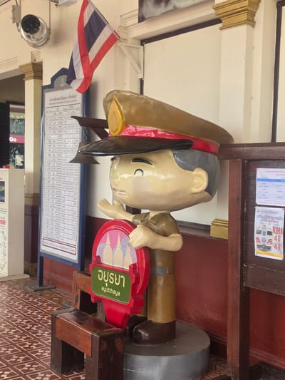 The image captures a scene within the Phra Nakhon Si Ayutthaya City Municipality, Thailand. The primary subject is a large, cartoon-like statue of a man, possibly representing a traveler or conductor. The statue is positioned next to a sign indicating the destination "Ayutthaya". Adjacent to the statue and sign is a schedule board with departure times of trains. The Thai flag is visible in the background, further indicating the location. The scene is set indoors, with architectural details suggesting a station or waiting area. The environment includes tiled flooring and wooden elements. The color palette is dominated by shades of brown, red, and beige.