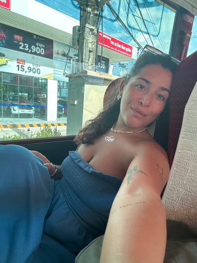 A young woman is taking a selfie while sitting in a vehicle. She has long, dark hair and is wearing a blue strapless top. She has a necklace with a name pendant and a pearl bracelet on her wrist. There are tattoos on her upper arm and shoulder. Through the window, a street scene in Kampot, Cambodia is visible, including billboards advertising Toyota cars with prices in US dollars. The weather appears to be clear and sunny. The overall mood seems relaxed and casual.