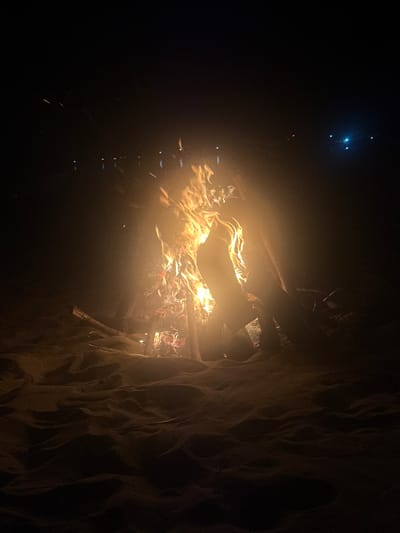 A crackling bonfire burns brightly in the night, illuminating stacked logs and sending embers into the darkness. To the right, a textured, possibly sandy or rocky structure looms, partially obscuring the fire. Distant lights glimmer faintly in the upper left corner, contrasting with the intense heat and glow of the flames. The fire casts long shadows, enhancing the depth and mystery of the scene.