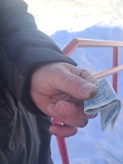 A close-up, outdoor shot in Bolshaya Elhovka, Russia, depicts an adult right hand holding a folded banknote. The hand, exhibiting visible dirt or grease on the fingertips and unmanicured nails, is partially covered by a dark, possibly winter jacket sleeve. The banknote, identified as a light blue and white Russian 50 Ruble note, is held between the thumb and index finger. In the mid-ground, a pink/red metal railing or pipe is present. The background is dominated by snow-covered ground, indicating a winter environment under bright, possibly overcast, daylight conditions.