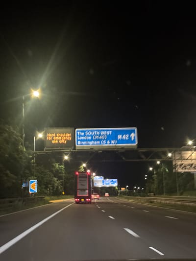 The image captures a nighttime scene on a multi-lane motorway in the Metropolitan Borough of Solihull, United Kingdom, viewed from a vehicle. The road is well-lit by numerous streetlights on overhead gantries, contrasting with the dark night sky.
In the foreground and mid-ground, prominent road signs guide traffic. A large blue overhead sign indicates directions for "The SOUTH WEST", "London (M40)", and "Birmingham (S&W)", with an arrow pointing straight ahead for "M42". Directly below this, an orange electronic variable message sign displays the text "Hard shoulder for emergency use only". Further along the hard shoulder, a smaller blue sign with a yellow telephone symbol and "SOS" is visible, indicating an emergency telephone point.
A red double-decker bus is seen from the rear, occupying one of the lanes, with its taillights and brake lights illuminated. The road features clear white lane markings and a solid white line separating the hard shoulder. Trees and dark foliage line the sides of the motorway. The scene depicts active nighttime travel on a busy, illuminated highway under clear weather conditions.