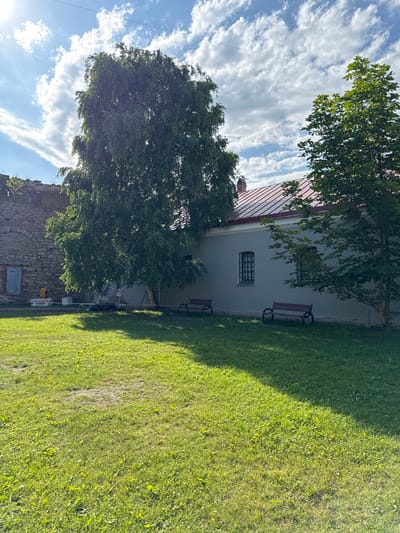 The image depicts an outdoor scene in Posyolok Imeni Morozova, Russia, on a sunny day with scattered clouds. The foreground is dominated by a lush green lawn, partially shaded by a large tree. To the left, a section of an old stone wall is visible, suggesting a historical setting.
Behind the stone wall and tree, a light-colored building with a red roof stands. The building has at least two windows with dark, barred frames. In front of the building, there are two wooden benches placed on a paved walkway. Another larger tree partially obscures the building.
In the lower right portion of the frame, a shadow is cast across the lawn, indicating the sun is at an angle, likely late afternoon or early morning. The sky is bright blue with wispy white clouds, contributing to the overall bright and airy feel of the image. There are no people clearly visible in the frame, and the scene appears peaceful and serene. The overall impression is that of a historical park or garden area on a pleasant day.