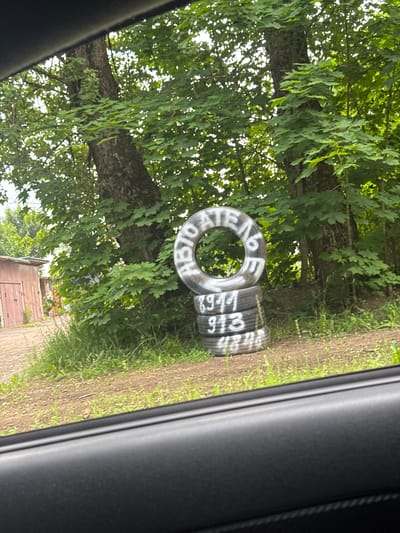 The image shows a stack of car tires on the side of a dirt path with a background of trees and foliage. The tires are painted with white lettering. One tire, lying flat on its side, has the word "АВТО" (AUTO) painted around its rim, with "ПН" (PN) visible above it and other letters and numbers obscured. Another tire, stacked on top of it, has the numbers "9168" painted on its sidewall. The scene appears to be outdoors in a rural or semi-rural setting, possibly in Kuzmolovskiy, Russia, given the provided location context. The lighting suggests it is daytime, with diffused sunlight filtering through the dense green leaves of the trees. The overall atmosphere is one of stillness and perhaps abandonment, with the tires possibly being discarded or stored. The image is taken from inside a vehicle, as indicated by the dark, curved frame on the right side of the picture, which could be a car door or window frame.