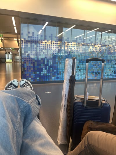 Here is a description of the image:
The image depicts the lower half of a person sitting, presumably in a transit waiting area. They are wearing blue jeans and black and white sneakers, which are positioned at the bottom of the frame. To the right, next to the person, is a blue suitcase. Resting on top of the suitcase is a brown bag. An umbrella wrapped in plastic is placed on the floor next to the suitcase. The background features a wall with a geometric pattern of blue squares. Through the large window, elements of the building's interior can be seen. The text on the plastic reads, in part: "Quantum dots exhibit discrete...spectra... tunable optical properties... infrared and terahertz regimes." The image location is Fischamend, Austria.