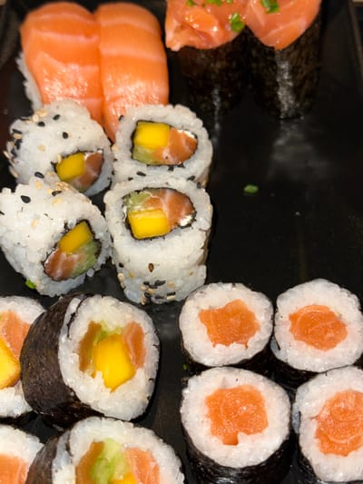 A close-up shot shows an assortment of sushi, including salmon nigiri with vibrant orange fish slices. Additionally, there are several varieties of sushi rolls. Some rolls are filled with salmon, mango, and avocado, coated in white rice and black sesame seeds. Others are simple salmon rolls wrapped with seaweed. The arrangement showcases a visually appealing and diverse sushi platter.