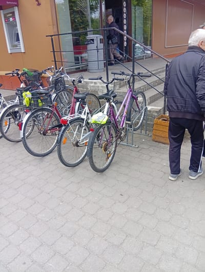 The image depicts an outdoor scene in Kuršėnai, Lithuania, in front of a building that houses an ATM. Several bicycles are parked in front of the building. Two men are visible in the foreground, one walking away from the camera and another with his back to the camera. The man with his back to the camera is wearing a dark jacket and dark pants, with athletic shoes. The other man, walking away, is wearing a dark jacket and lighter pants. The ATM is labeled "Luminor" and is mounted on the building's exterior wall. The ground is paved with interlocking gray stones. A trash receptacle is visible near the building entrance.
