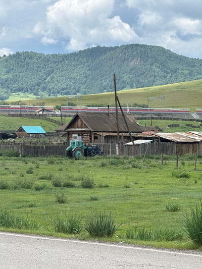 The image depicts a rural village scene in Kazanovka, Russia, under a cloudy sky. In the foreground, a paved road runs horizontally, bordering a grassy field. The field contains scattered green bushes and tall grasses. Further back, a wooden fence encloses a residential area. Within the enclosure, a rustic wooden house with a weathered roof stands prominently. Adjacent to the house is a green tractor with a front loader. Another smaller building with a bright blue roof is visible to the left, also enclosed by the wooden fence. In the mid-ground, a train with red and white stripes travels on elevated tracks. The background is dominated by a lush, green, tree-covered mountain slope.