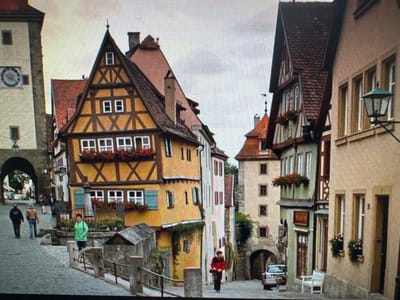 This image depicts a quaint European street scene, likely in Ellwangen, Germany, with its characteristic half-timbered buildings. The perspective is from above, looking down at the street, and the image is rotated, making it appear upside down.
The buildings themselves are the primary subjects, with their colorful facades, steep roofs, and numerous windows, many adorned with flower boxes filled with red blooms. The architectural style suggests a historical European town. One building, in particular, stands out with its prominent yellow and brown half-timbered design, featuring a large triangular gable.
There are a few people visible, scattered throughout the scene. One person, wearing a red top, is visible near the top of the image, appearing to be walking. Another individual, dressed in green, is near a window on a higher floor. Further down, a person in black is standing beside a tall, dark object, possibly a signpost or statue. The presence of these individuals suggests a normal day in a populated area.
Notable details include the presence of a large clock on one of the buildings on the right side of the image. The sky appears overcast, suggesting a cloudy day. The overall atmosphere of the scene is one of historical charm and everyday life in a European town. No visible text is discernible in the image.