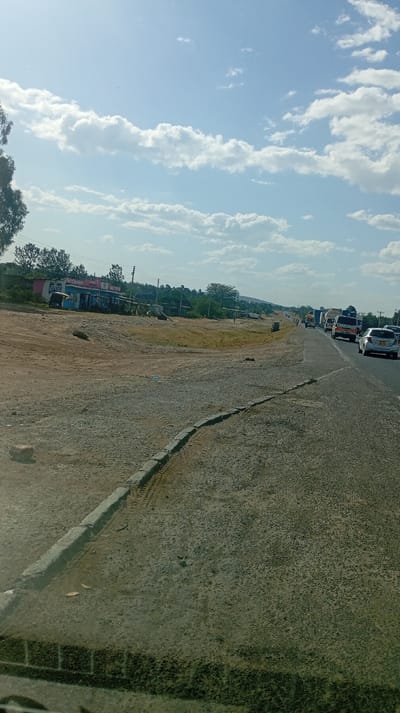 An outdoor scene shows a road filled with vehicles under a bright, partly cloudy sky. The shoulder of the road is dirt-covered. Along the roadside, there are sparse buildings and patches of green vegetation. Distant hills can be seen on the horizon. The perspective is from the inside of a car, looking out onto the scene.
