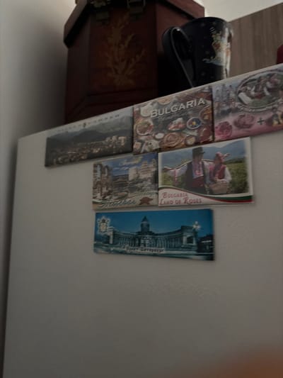 The image displays a white refrigerator door adorned with several rectangular postcards. The postcards are arranged in a grid formation, with some overlapping. The top left postcard features a panoramic view of a city nestled in a mountainous landscape. To its right, a postcard titled "BULGARIA" showcases a collage of food items. Below the "BULGARIA" postcard, another postcard depicts a couple in traditional Bulgarian attire standing in a field, with the text "BULGARIA LAND OF ROSES" at the bottom. A postcard to the left of this one shows ruins of a building and the word "Brezhtar" in Cyrillic script. The bottom postcard features a grand architectural building with columns and a dome, and the text "Pteresburg" which likely refers to St. Petersburg. Above the postcards, part of a dark wooden chest with ornate carvings is visible, along with a dark blue mug with a celestial pattern. The scene appears to be an interior setting, likely a kitchen, with the refrigerator being the primary subject. There is no indication of the location being Dromolaxia - Meneou Municipality, Cyprus.