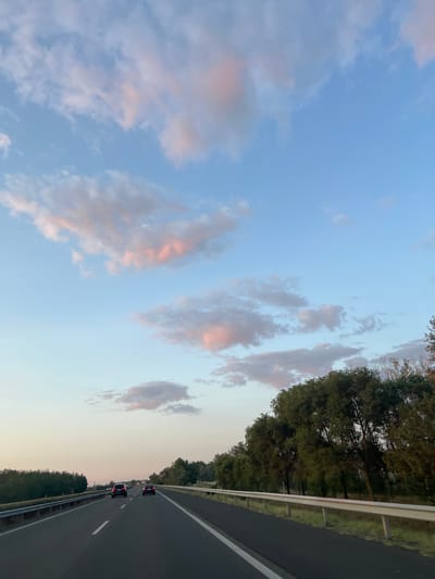 The image depicts a highway scene at dusk, with a clear sky transitioning from blue to a pale yellow near the horizon. Several pink-tinged clouds are scattered across the sky. The road, with white lane markings, stretches forward, leading towards two dark-colored cars in the distance. To the right of the road, a line of dense green trees forms a natural barrier, with a white guardrail running alongside it. The left side of the road also shows trees and vegetation. The overall atmosphere suggests travel on a highway in Balástya, Hungary, during the evening.