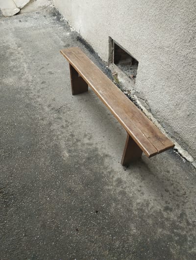 A rustic wooden bench is positioned on an asphalt surface alongside a textured, light-colored wall. To the left of the bench, the asphalt appears worn and somewhat mottled. To the right, against the wall, there is a dark, triangular opening revealing the brickwork behind it. The scene is set in an urban environment, specifically in Vinkivtsi, Ukraine, under natural lighting conditions. There are no people or animals visible in the image.