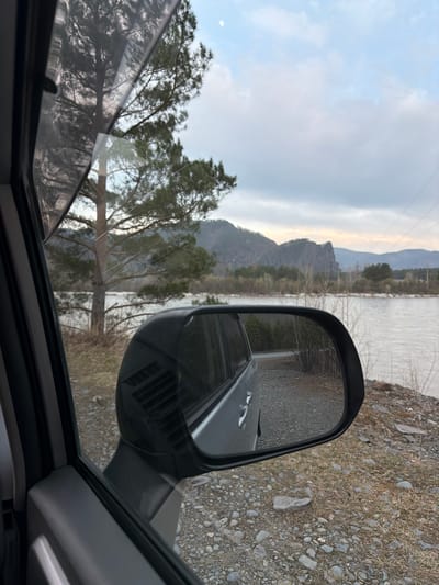 The image presents a view from inside a vehicle, looking out its side window and into the attached side mirror. The scene depicts a tranquil, rural landscape in Abaza, Russia.
Through the vehicle's window, a wide body of water, likely a river or large stream, dominates the midground, running parallel to the vehicle's apparent path. Beyond the water, distant mountains or hills rise, their contours softened by a hazy, partly cloudy sky. The horizon shows faint warmer tones, suggesting either early morning or late afternoon light. Bare trees line the riverbank, and a larger, leafy tree stands closer to the vehicle on the left side of the frame. Power lines are subtly visible against the sky and mountains.
The prominent dark side mirror on the right reflects the immediate surroundings of the vehicle: the side of the car, a dirt and gravel track or road, and the stony, sparsely vegetated ground adjacent to it. The ground outside the vehicle is a mix of pebbles, rocks, and dry grass. The window itself shows some smudges and reflections. No people are visible, and no specific activity is taking place beyond observing the scenery from the stationary or slowly moving vehicle. There is no visible text in the image.