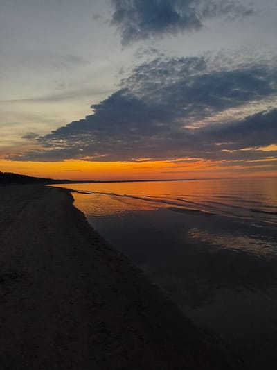 This is a landscape photograph of a beach at sunset in Apšuciems, Latvia. The image captures the Baltic Sea meeting a sandy shore. The sky is a gradient of colors, with a bright orange band near the horizon transitioning to darker blues and grays towards the top, punctuated by dramatic cloud formations. The calm sea reflects the vibrant colors of the sunset. A dark, forested coastline is visible along the left edge of the image, forming a silhouette against the bright sky. The sand in the foreground is dark and appears to have footprints. The overall atmosphere is serene and natural.