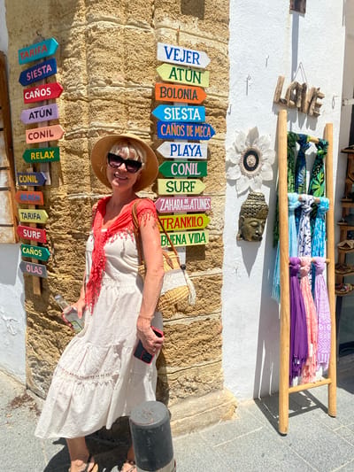 In Conil de la Frontera, Spain, a woman with blonde hair and sunglasses, wearing a white dress and a straw hat, stands against a textured stone wall. She holds a bottle in her left hand and a smartphone in her right. A woven straw bag hangs on her shoulder. The wall is adorned with colorful directional signs pointing to various locations including "Tarifa," "Siesta," "Caños," "Atún," "Conil," "Relax," "Playa," and "Surf." To the right, a white wall displays a driftwood "LOVE" sign and a decorative flower. A wooden rack holds several brightly colored scarves. A small golden Buddha head is mounted on the wall. The ground is paved with stone tiles, and a dark cylindrical object is visible in the foreground.