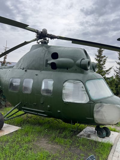 The image features a static display of a green Mil Mi-8 helicopter, likely in a park or outdoor museum setting. The helicopter's cockpit windows are prominent, and the main rotor blades are visible against a cloudy sky. The aircraft is situated on a concrete platform, surrounded by green grass and foliage. Given the mention of Введенская Слобода, Russia, it can be assumed that this display is located within or near that city.