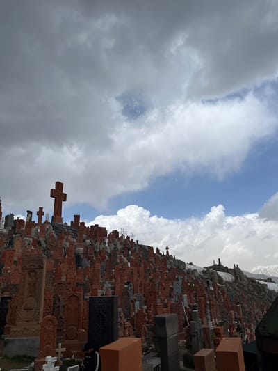 The image shows the cemetery in Hartagyugh, Armenia, with a dense collection of khachkars (Armenian cross-stones) covering a hillside. The khachkars vary in size and color, with many being a reddish-brown hue. There are snow-covered mountains in the background behind the crosses. The sky is filled with a mix of dark and white clouds, suggesting changing weather conditions.