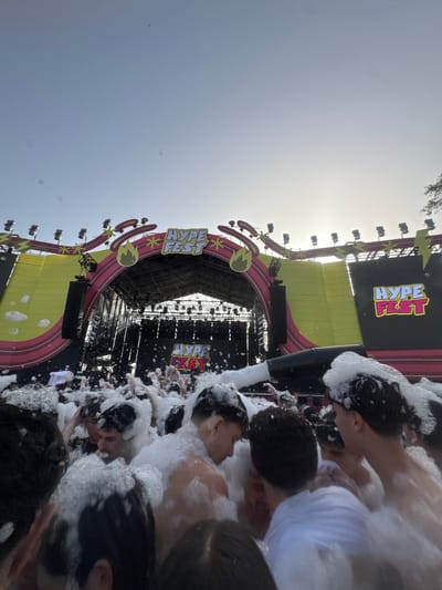 The image shows a crowd of people covered in foam at a "Hype Fest" event, presumably in Grau I Platja, Spain, given the geotag. The event features a large stage with the "Hype Fest" logo prominently displayed. The sky is visible, indicating the event is taking place during the day. Foam is being sprayed onto the crowd, creating a party-like atmosphere. The crowd appears dense and engaged.